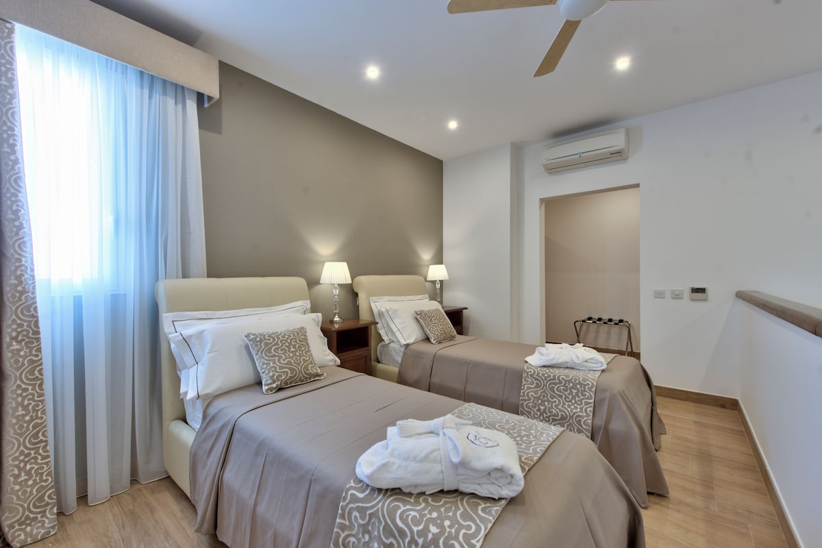 A hotel room with two beds, bedside lamps, a ceiling fan, an air conditioning unit, and a window with curtains.