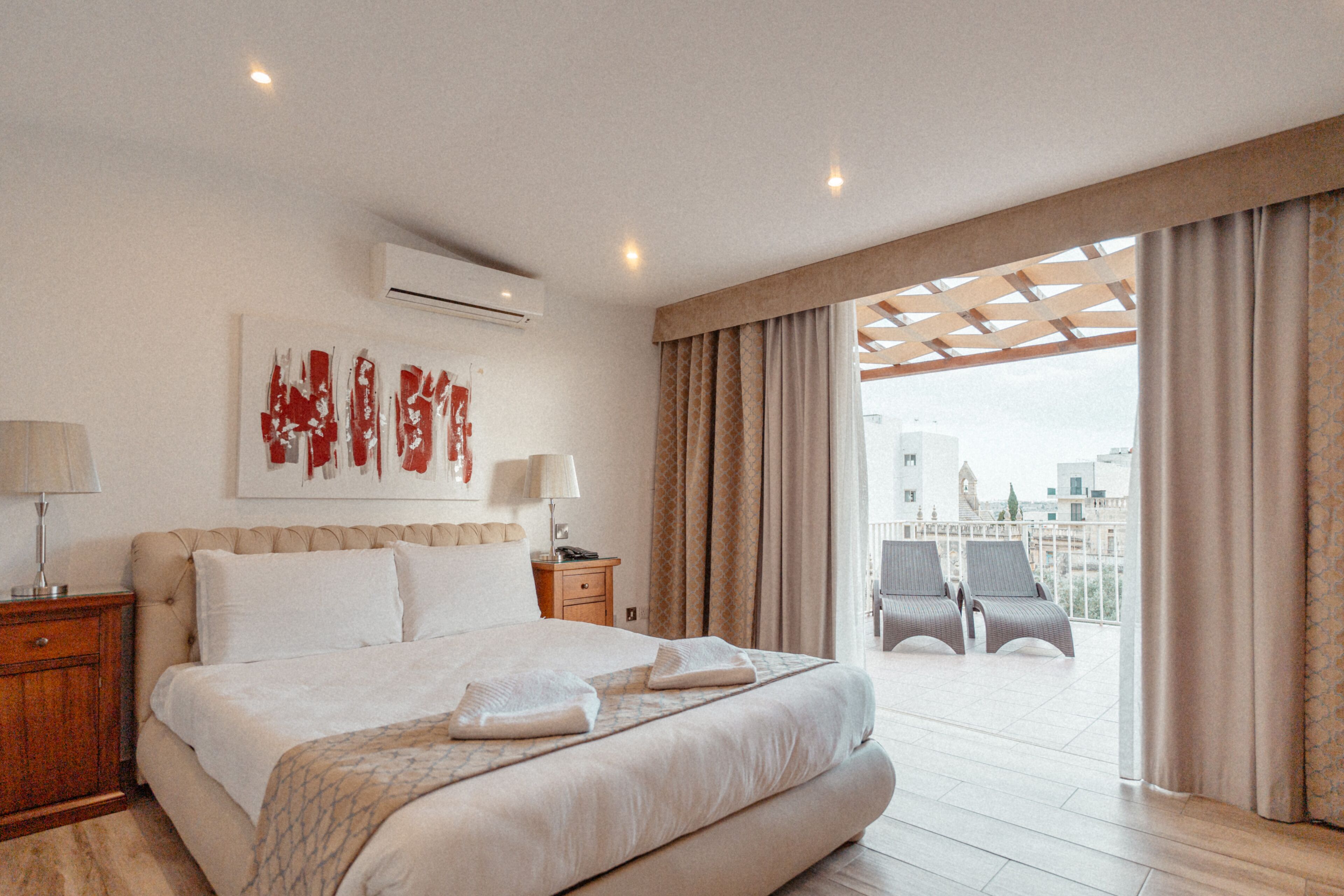 Habitación de hotel con una cama grande, una mesita de noche, una lámpara, aire acondicionado y vistas a la ciudad a través de amplias ventanas.
