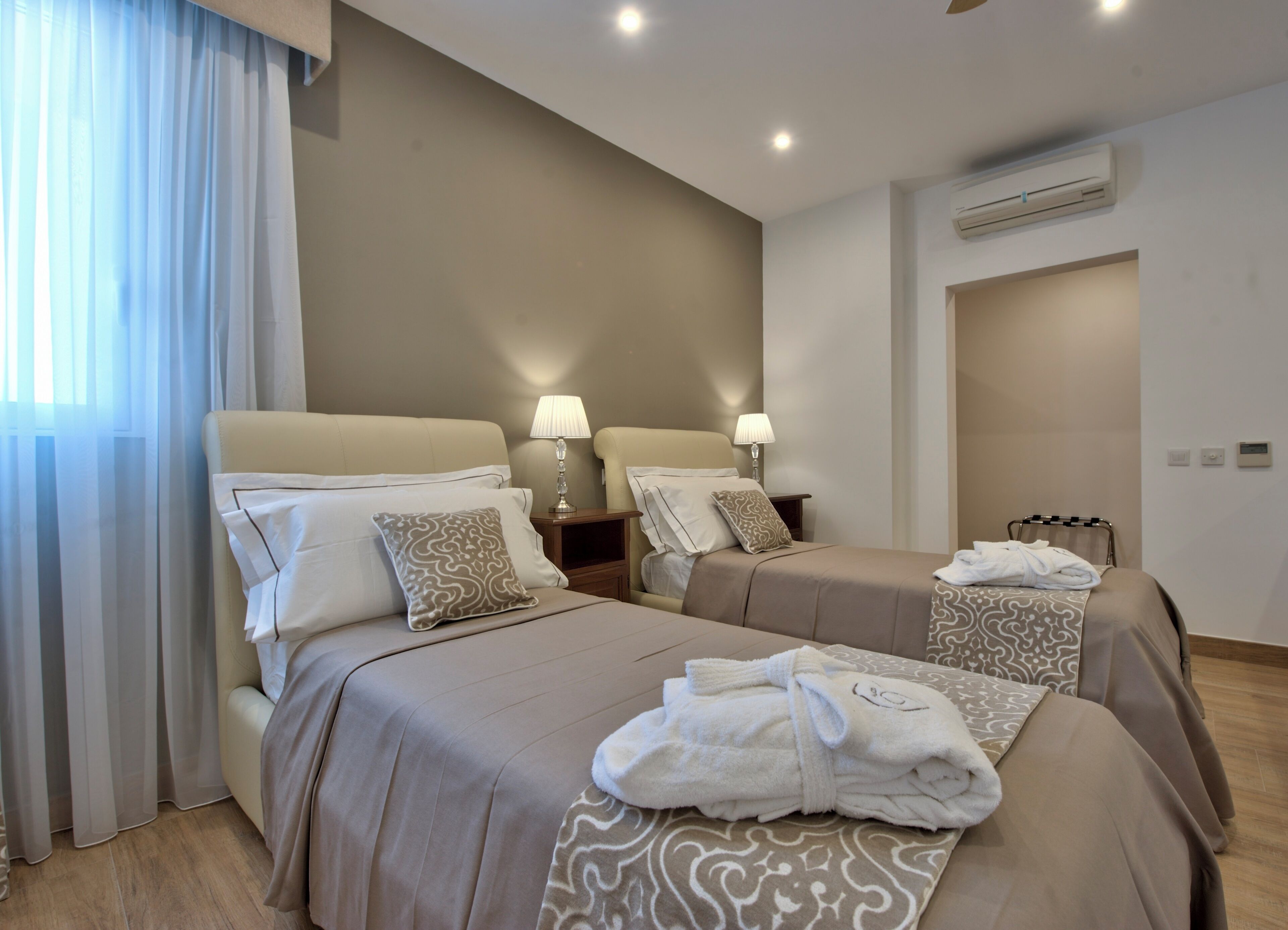 A hotel room with two beds, bedside lamps, an air conditioning unit, and a window with sheer curtains.