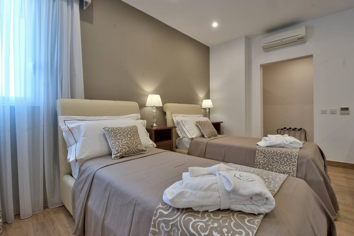 Una habitación de hotel con dos camas, lámparas de noche, un equipo de aire acondicionado y una ventana con cortinas traslúcidas.