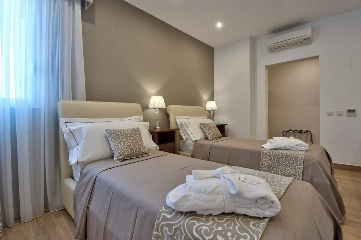 A hotel room with two beds, bedside lamps, an air conditioning unit, and a window with sheer curtains.