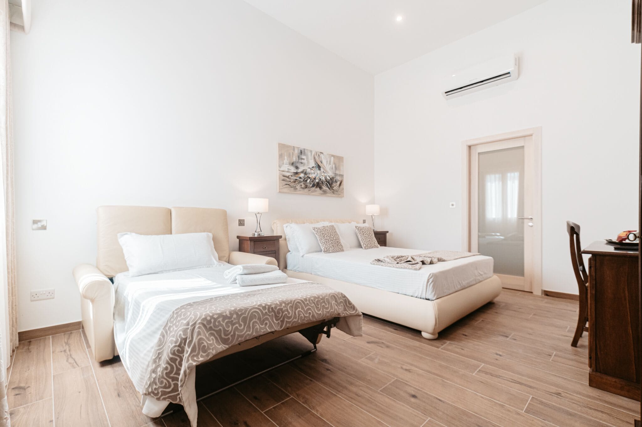 A hotel room with two beds, a wooden floor, a painting on the wall, and a door.