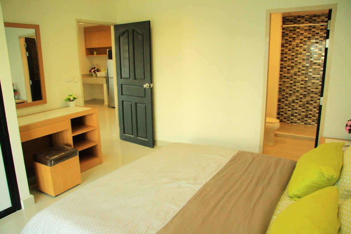 A bedroom with a bed, a wooden dresser, a mirror, and a bathroom visible through an open door.