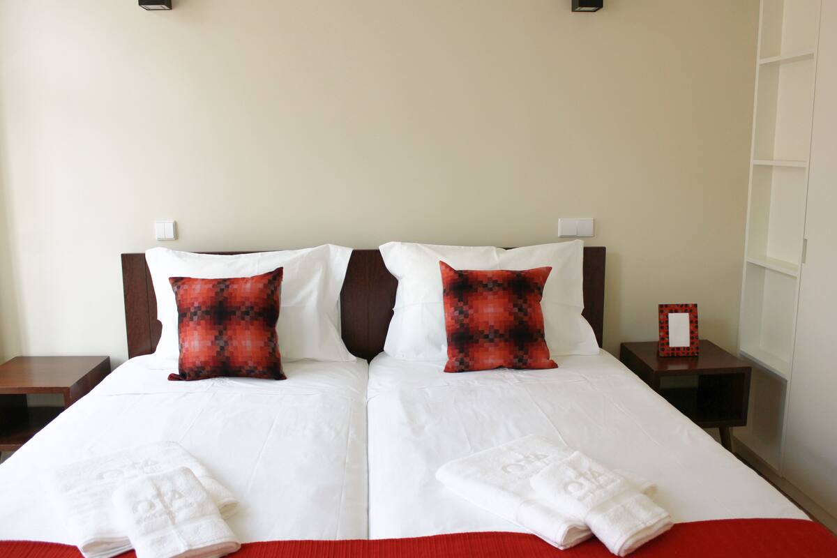 Un lit double avec du linge de lit blanc, deux coussins aux motifs rouges et noirs, et un plaid rouge.