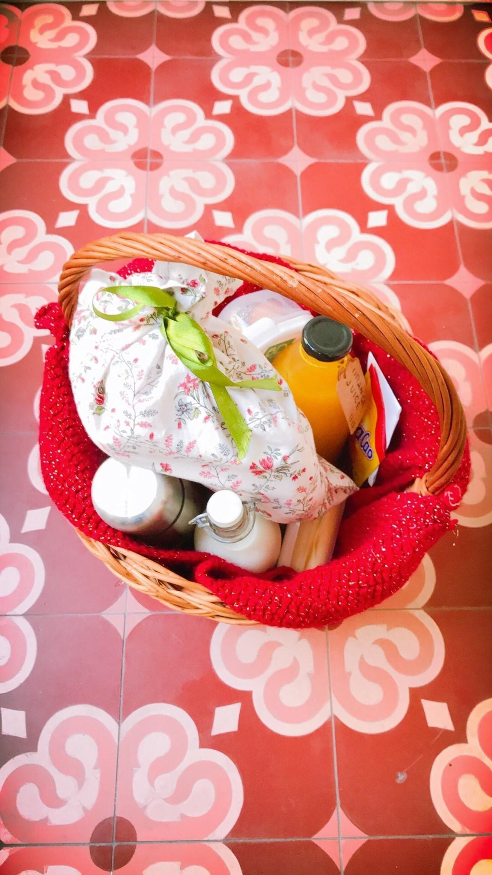 A wicker basket filled with various items, including a bottle of sauce, a jar, and a wrapped object, placed on a red patterned floor.