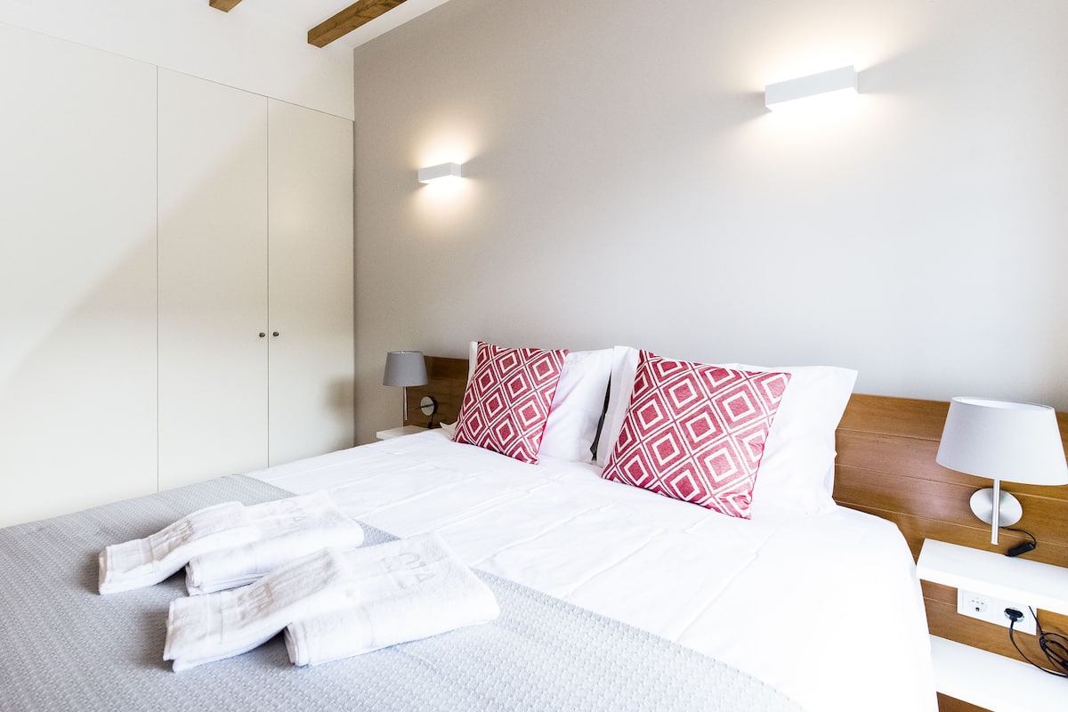 A neatly made bed with white bedding and red patterned pillows, a bedside table with a lamp, and a wall-mounted light fixture.