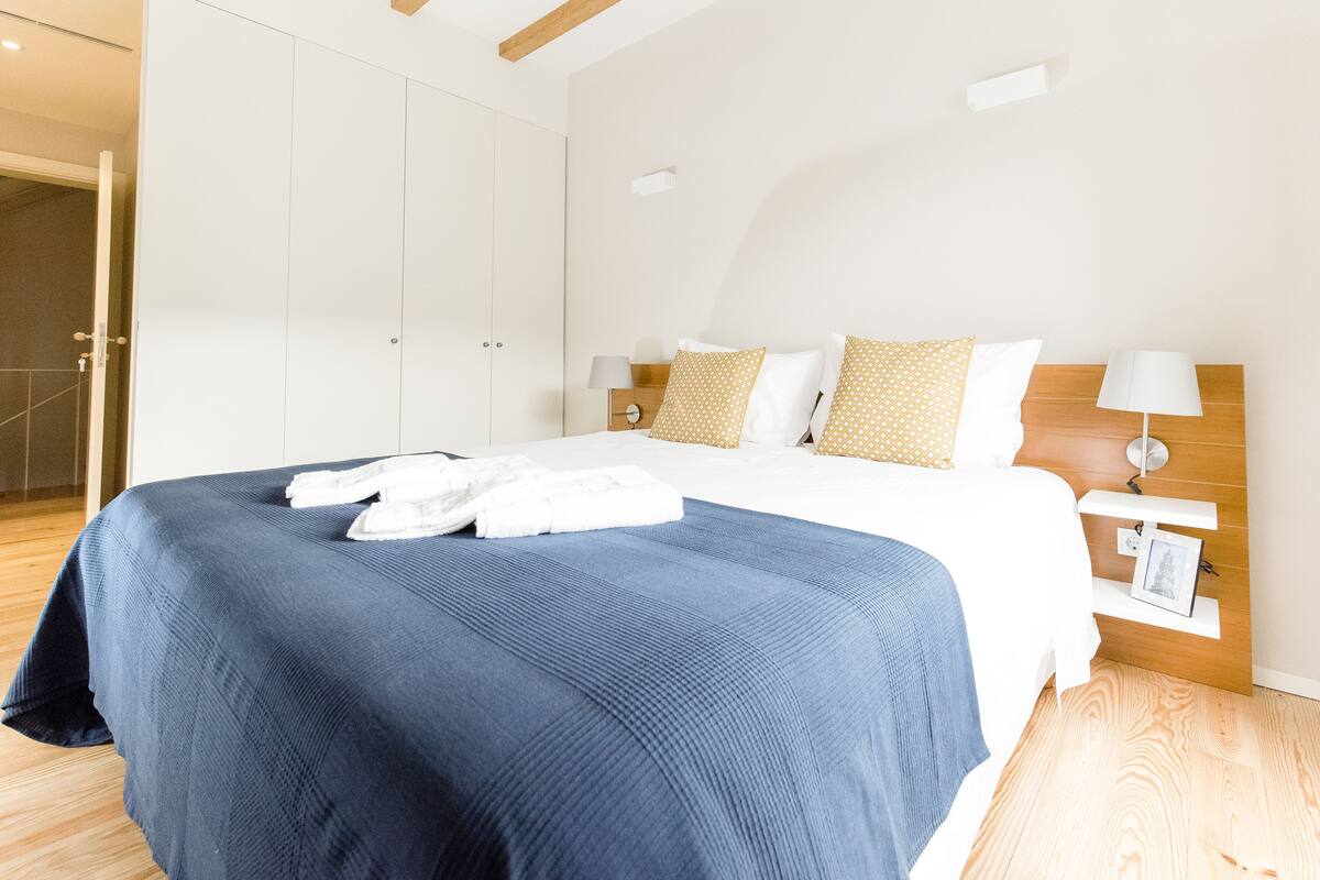 A neatly made bed with a blue bedspread and white linens, a wooden headboard, and a nightstand with a lamp.