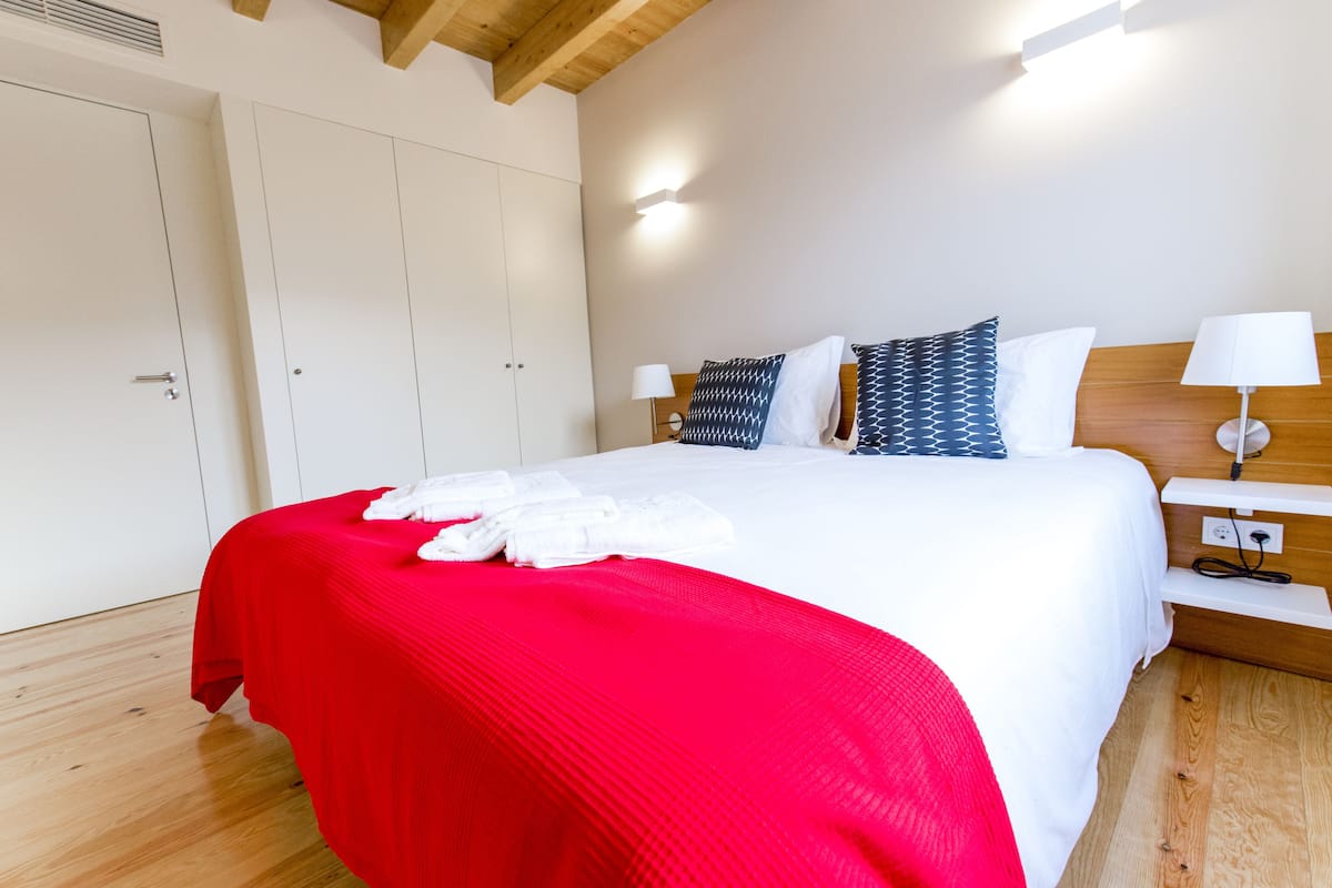A hotel room with a large bed, a red bedspread, white linens, a wooden headboard, two bedside lamps, and a white wardrobe.