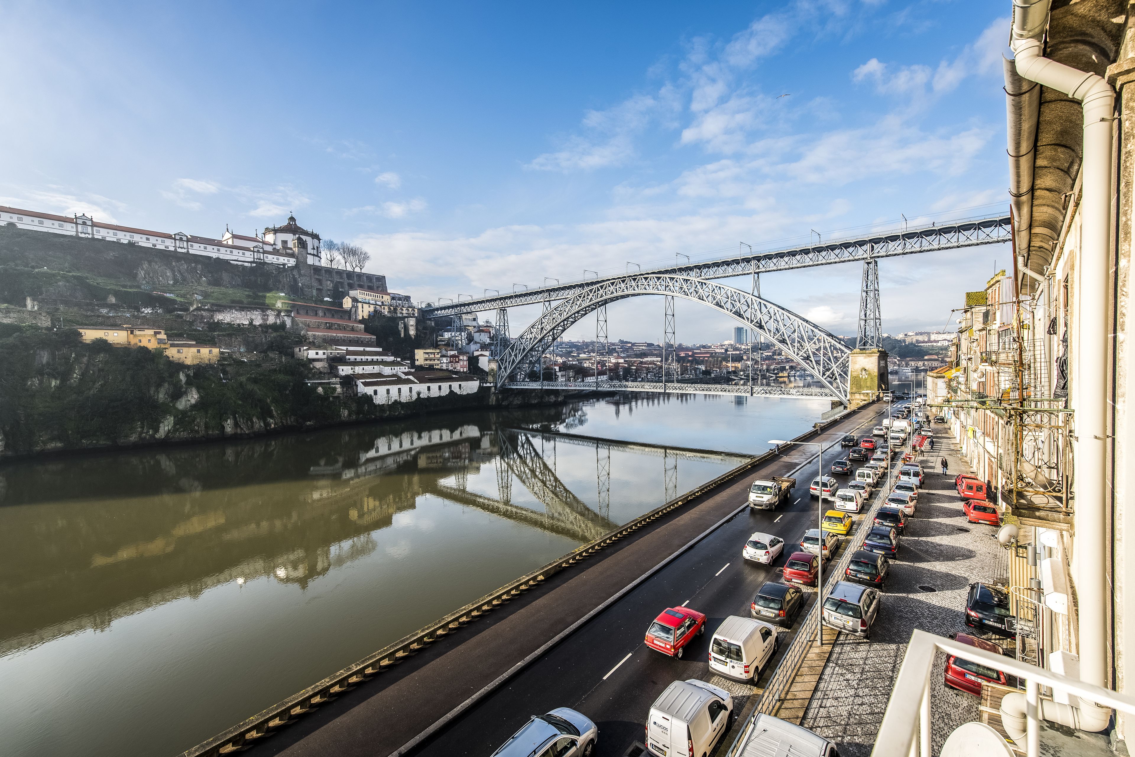 A busy urban street with a prominent bridge, a river, and a row of parked cars along the sidewalk.