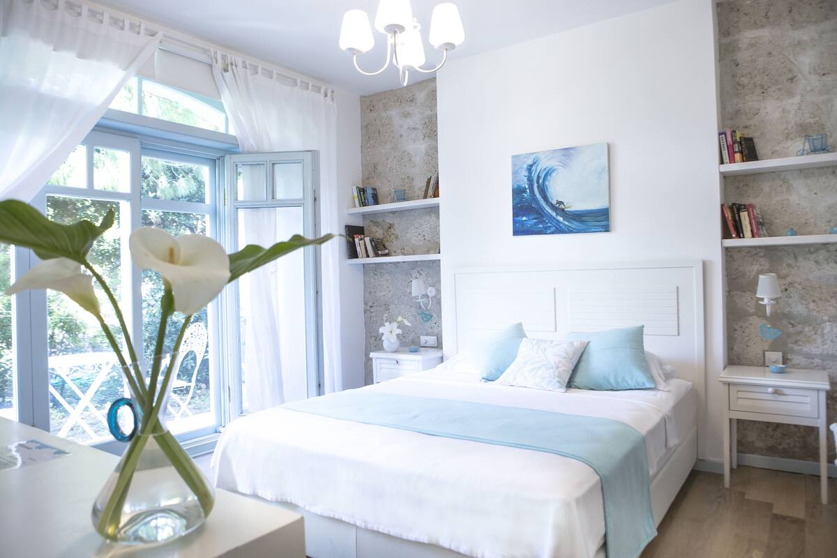 Ein Schlafzimmer mit einem Bett, Nachttischen, einem Fenster mit Vorhängen, einer Vase mit Blumen und einem Wandbild.