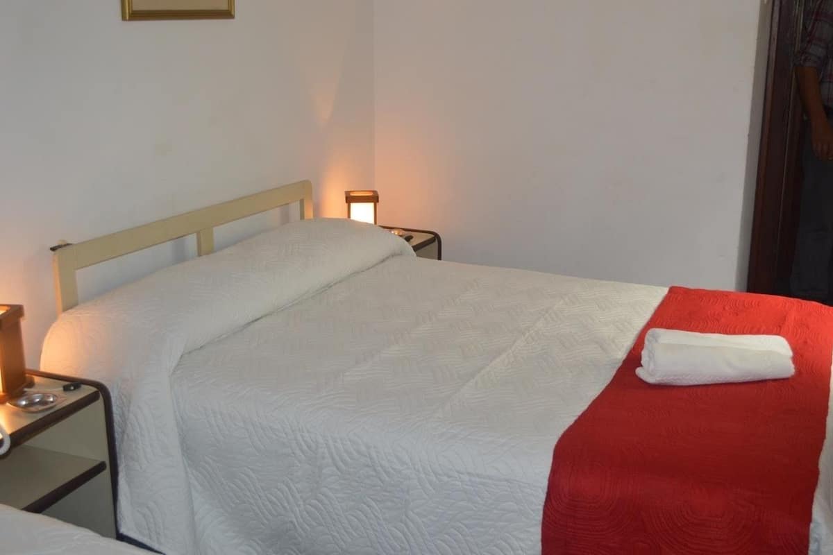 A neatly made bed with a white and red blanket, a small bedside table with a lamp, and a wall-mounted light fixture.