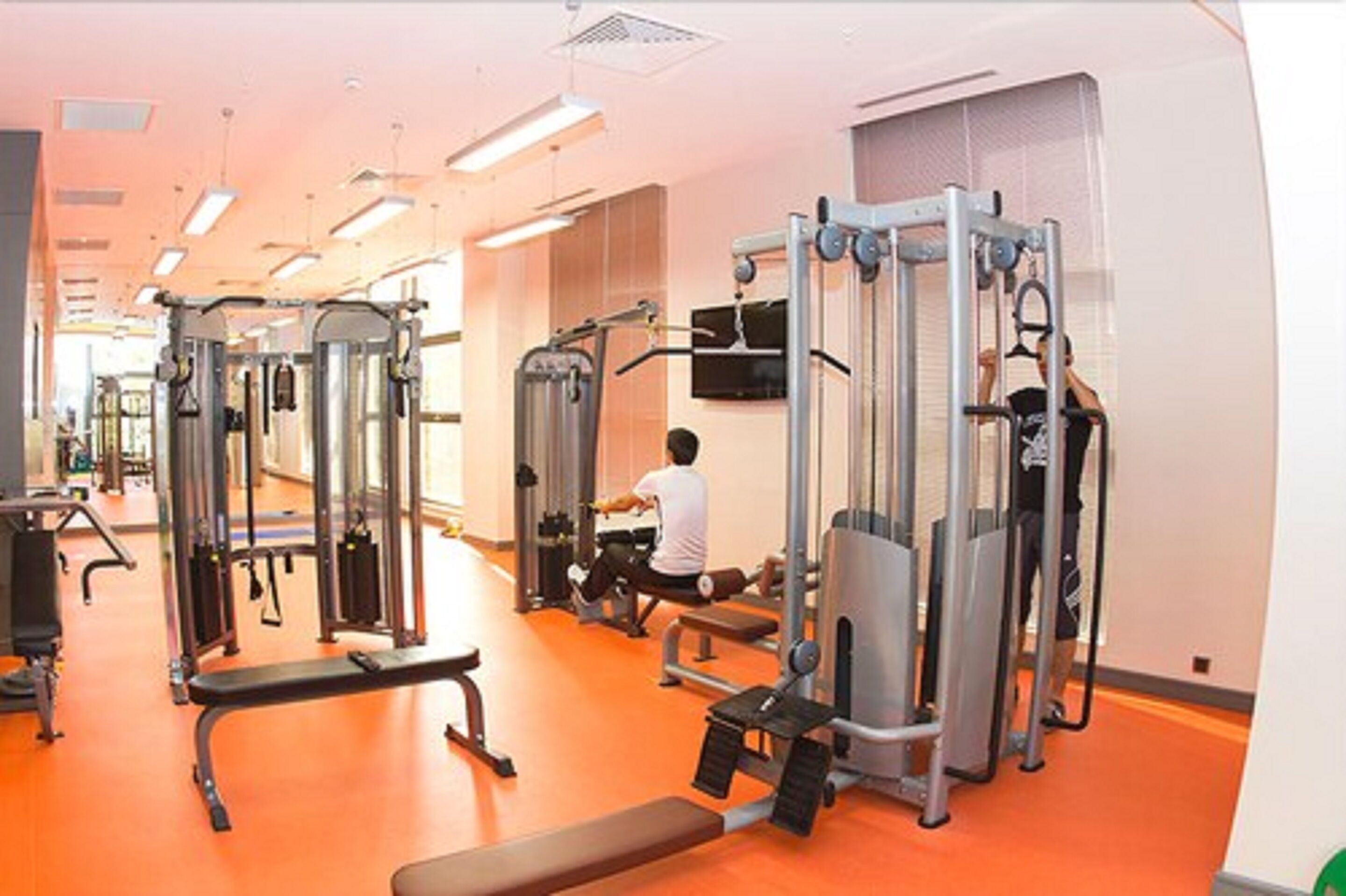 A gym with various exercise machines, a television mounted on the wall, and a person using a leg press machine.