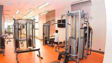 A gym with various exercise machines, a television mounted on the wall, and a person using a leg press machine.