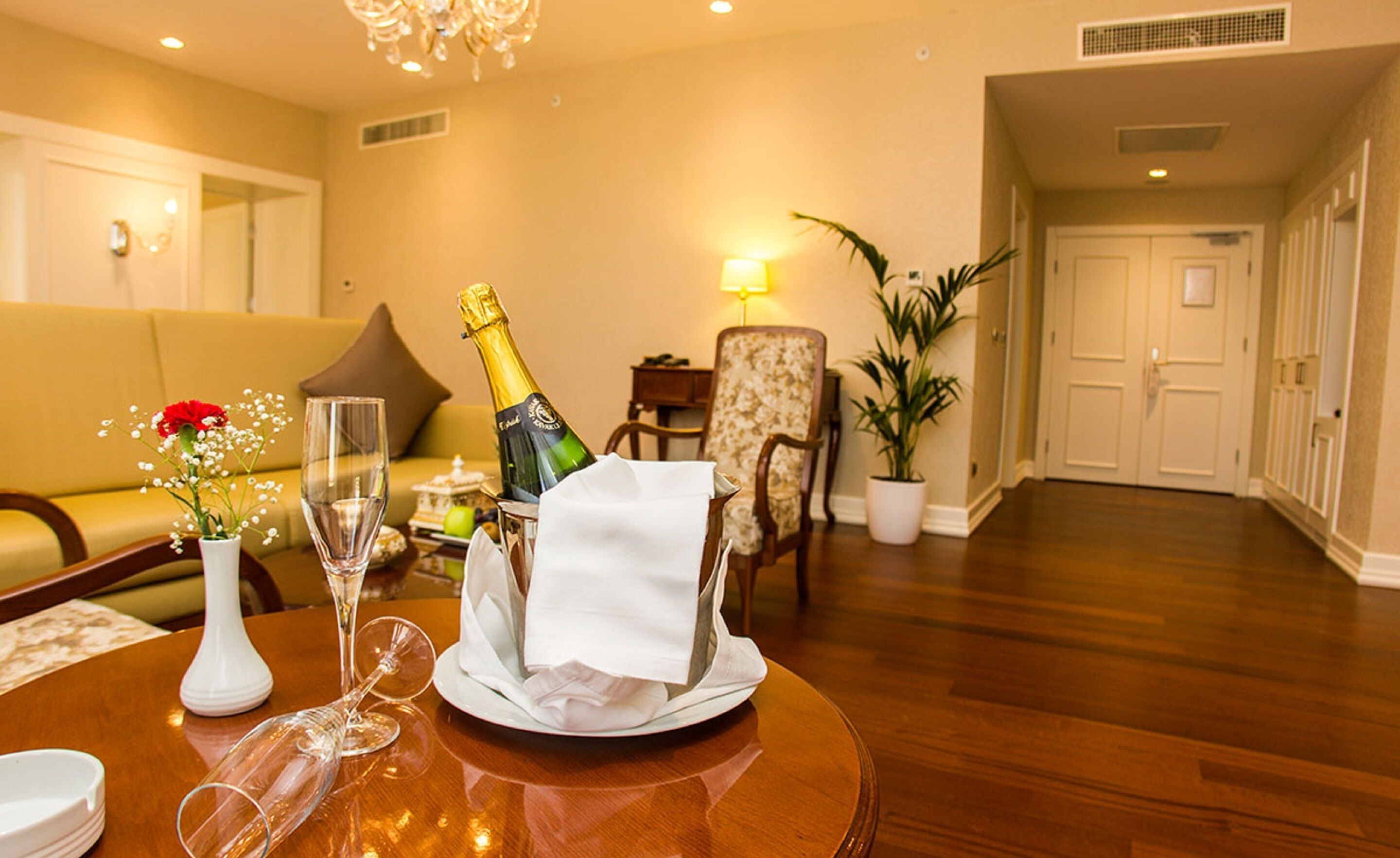 A well-lit living room with a wooden table set for a meal, a bottle of champagne, and a vase with flowers.
