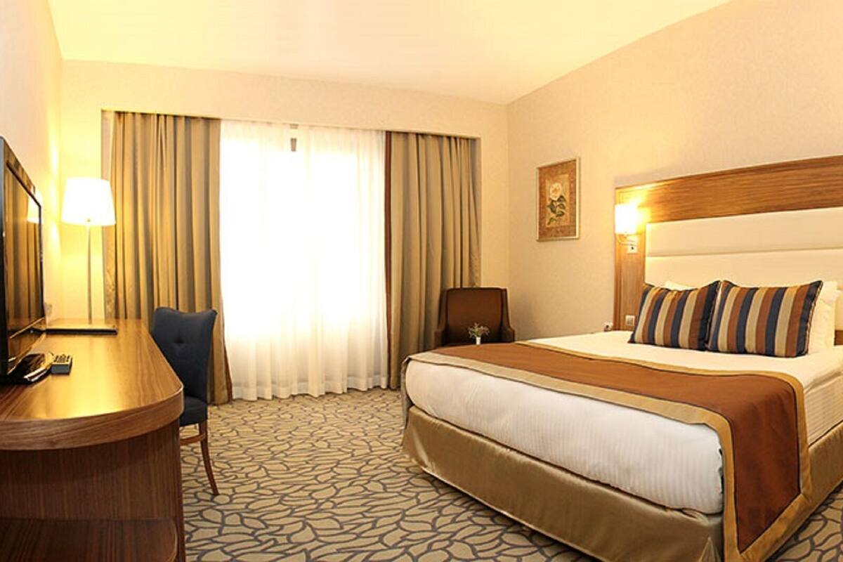 Habitación de hotel con una cama grande, un escritorio con computadora, una silla, una mesita con lámpara y un baño visible a través de una puerta abierta.