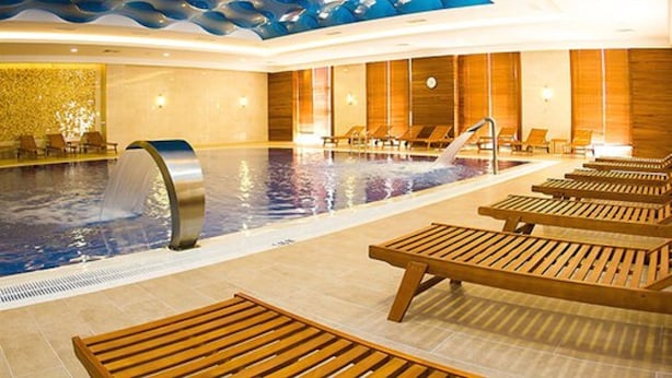 A modern indoor pool area with a wave-patterned ceiling, wooden loungers, and a central water feature.