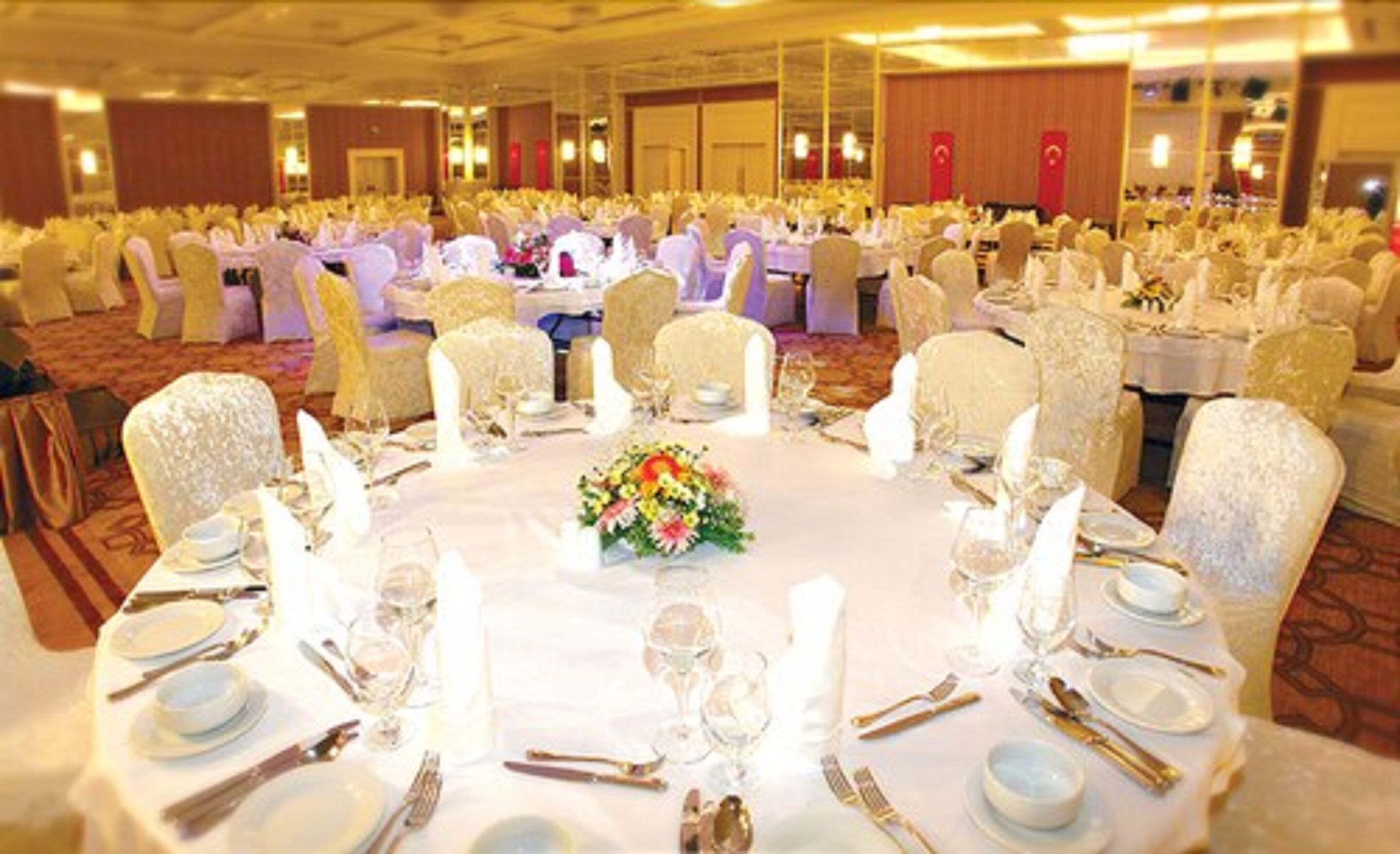 Un salón de banquetes con mesas redondas dispuestas para un evento formal, con manteles blancos, centros de mesa florales y cubiertos dorados.