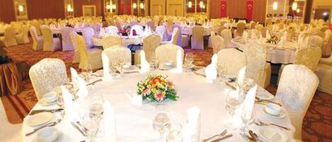 Un salón de banquetes con mesas redondas dispuestas para un evento formal, con manteles blancos, centros de mesa florales y cubiertos dorados.