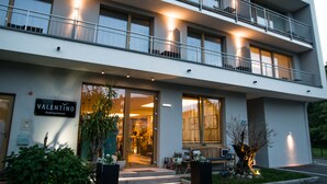 Un hotel moderno con un letrero que dice "Valentino Boutique Hotel". Cuenta con una estructura de dos pisos, balcones, ventanas amplias y una entrada acogedora con asientos al aire libre.
