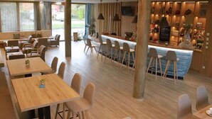 Un restaurante moderno con mesas y sillas de madera, una zona de bar con mostrador y amplios ventanales que ofrecen vistas al exterior.