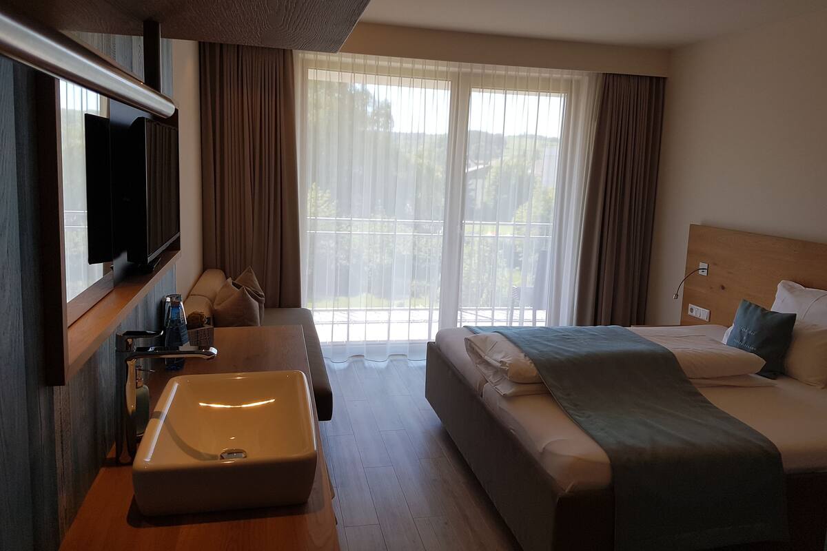 A hotel room with a bed, a sofa, a TV, a sink, and a balcony with curtains.