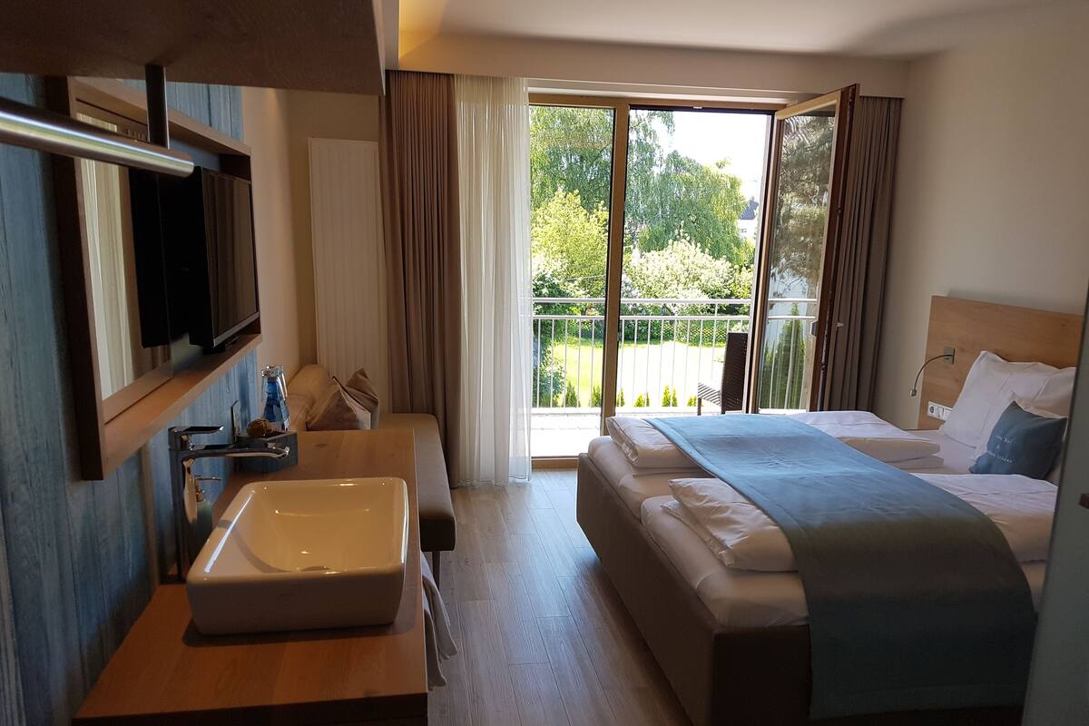 A hotel room with a large bed, a bedside table with a sink, a mirror, and a view of a balcony with greenery.