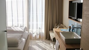 Habitación de hotel con cama, escritorio con lavabo, televisor y ventana grande con cortinas.