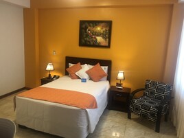Una habitación de hotel con cama, dos mesitas de noche con lámparas, una silla y un cuadro en la pared.