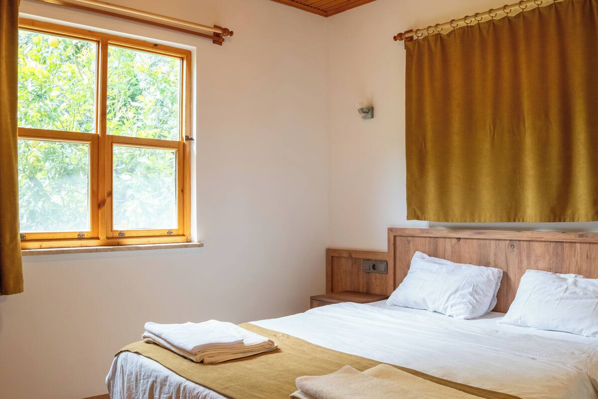 Ein Zimmer mit Bett, Fenster, Nachttisch und Vorhang.