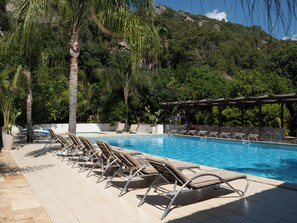 A pool area with lounge chairs and a pergola, surrounded by palm trees and a mountainous backdrop. - Cirali Hotel (Kemer)