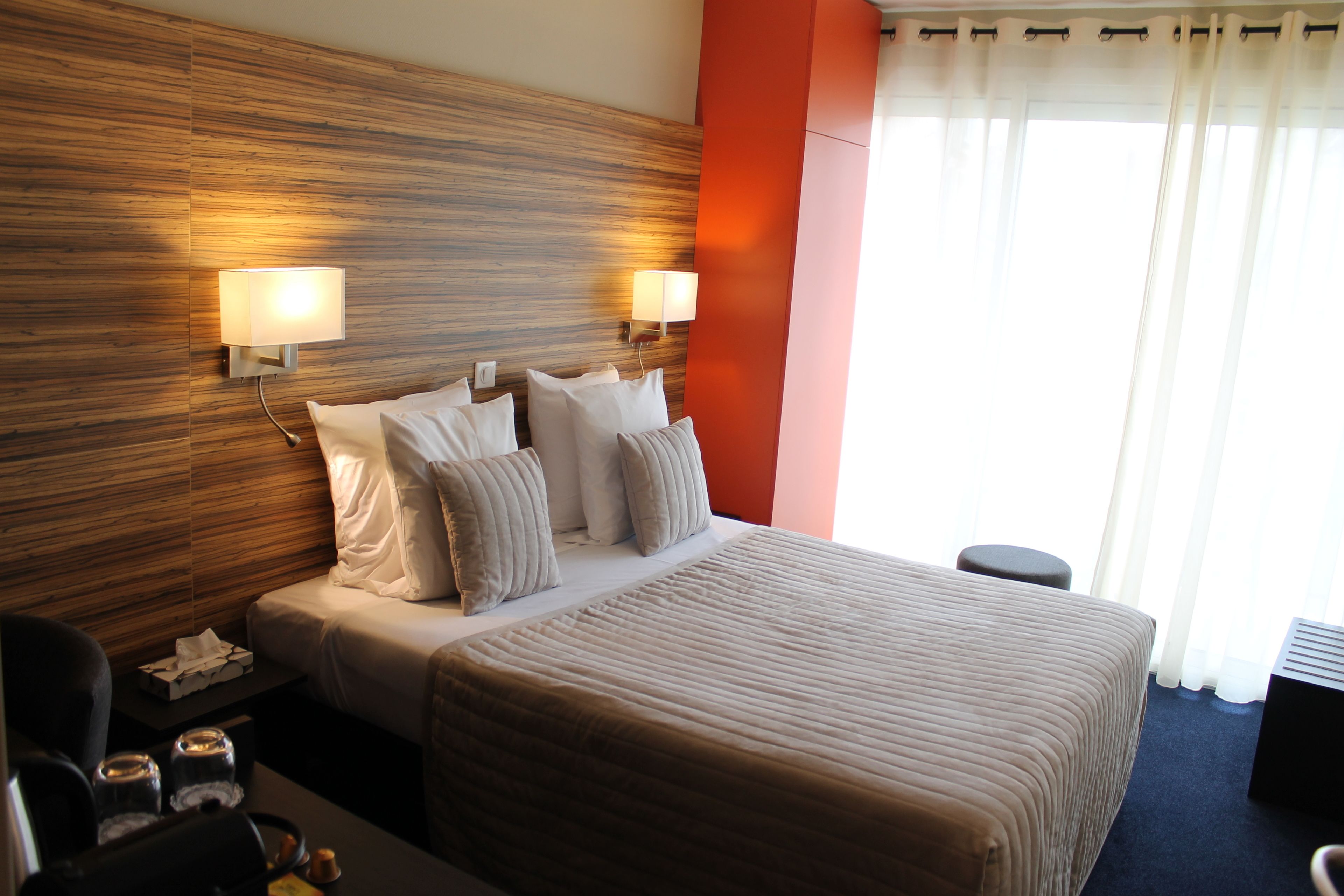 Habitación de hotel con una cama grande, dos lámparas de noche, un cabecero de madera, una pared de acento roja y una ventana con cortinas.
