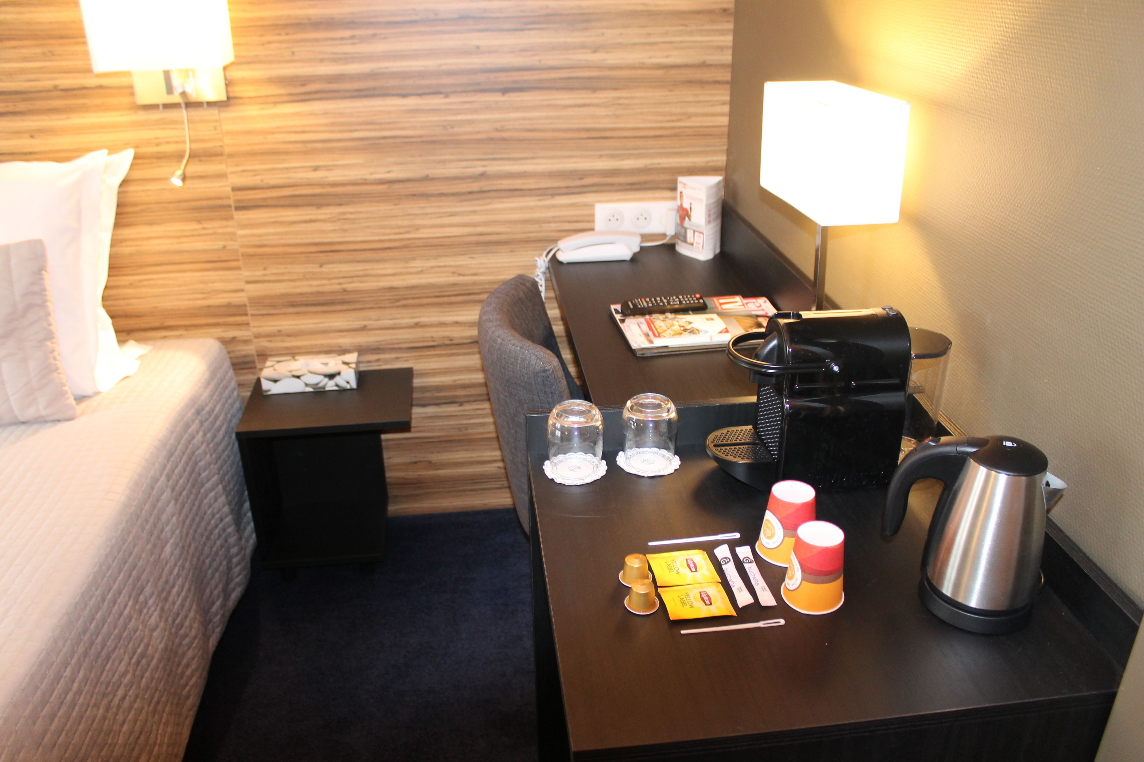 A hotel room with a bed, a desk with a coffee maker, cups, and a kettle.