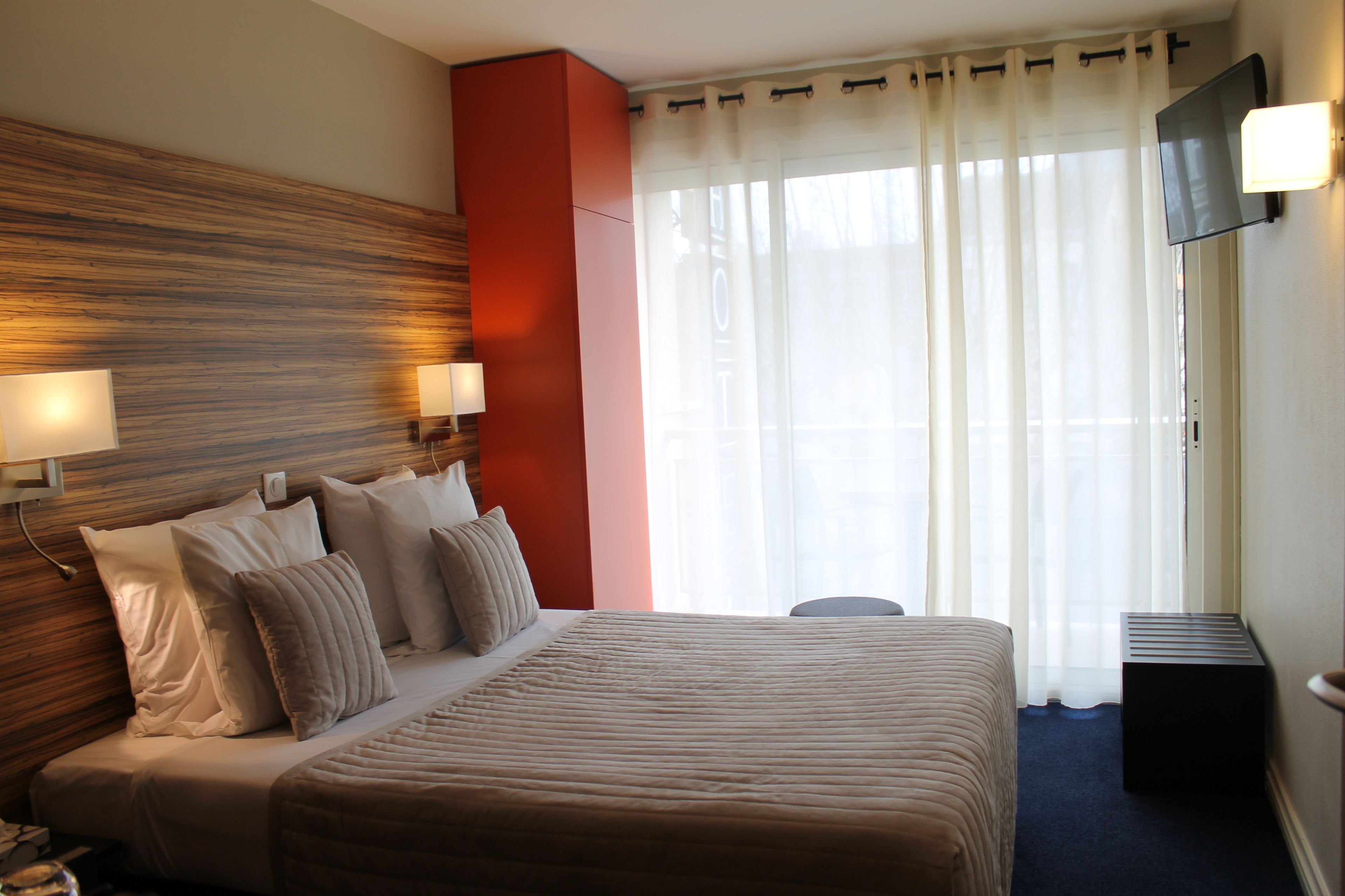 Ein Hotelzimmer mit einem großen Bett, einem an der Wand befestigten Fernseher und einem Fenster mit durchscheinenden Vorhängen.
