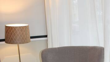 Ett hotellrum med ett träbord dukat för frukost, en grå fåtölj, en lampa och ett fönster med gardiner.