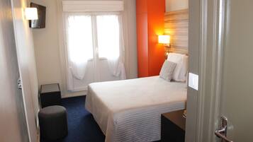 Habitación de hotel con una cama grande, una mesita de noche, una lámpara, una ventana con cortinas y una puerta.