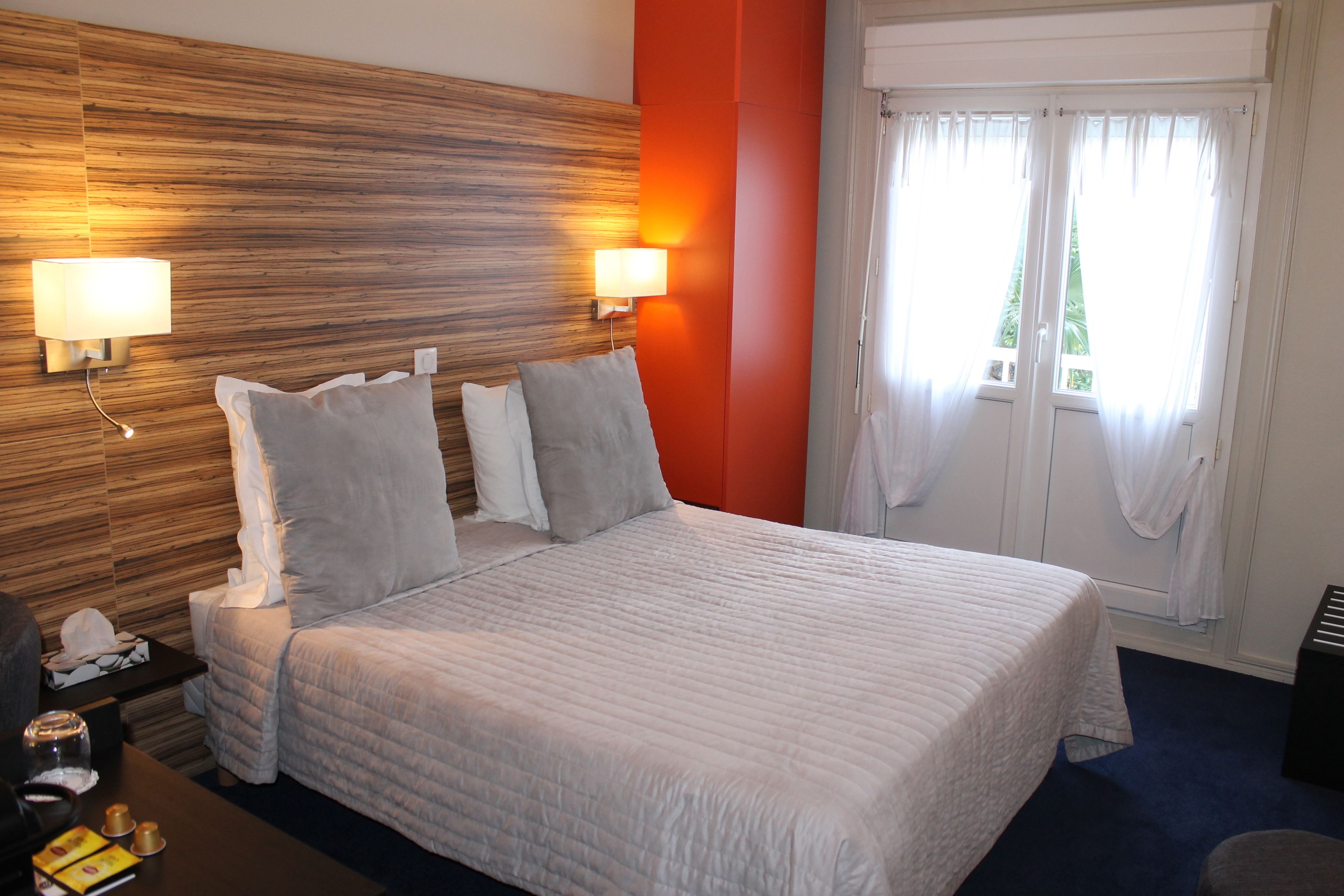 A hotel room with a large bed, two bedside lamps, a wooden headboard, a window with curtains, and a small table with a phone and a glass.