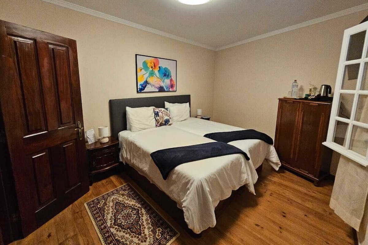 A bedroom with two beds, a wooden wardrobe, a nightstand, a decorative rug, and a painting on the wall.