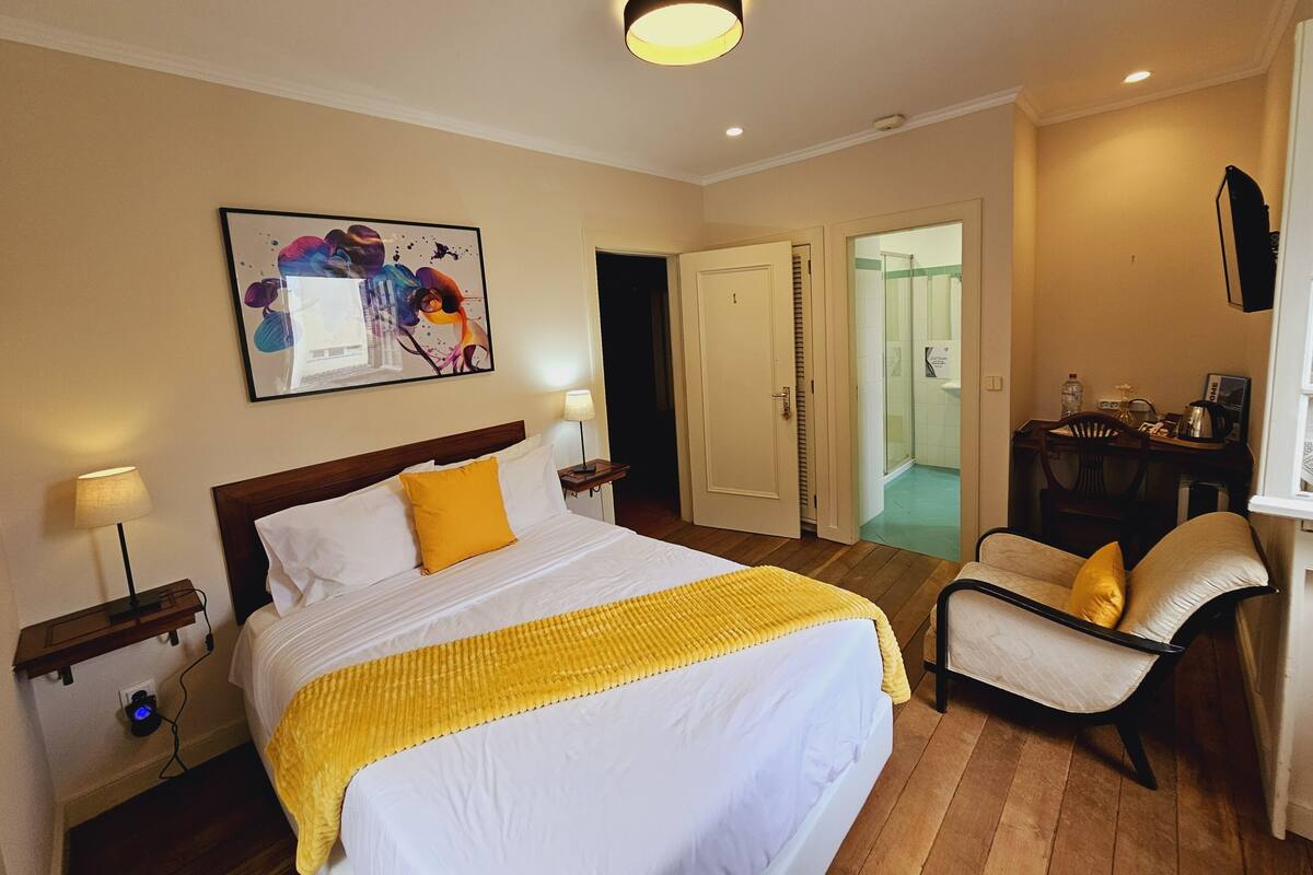 A hotel room with a large bed, a bedside table with a lamp, a chair, a desk with a chair, and a bathroom visible through an open door.