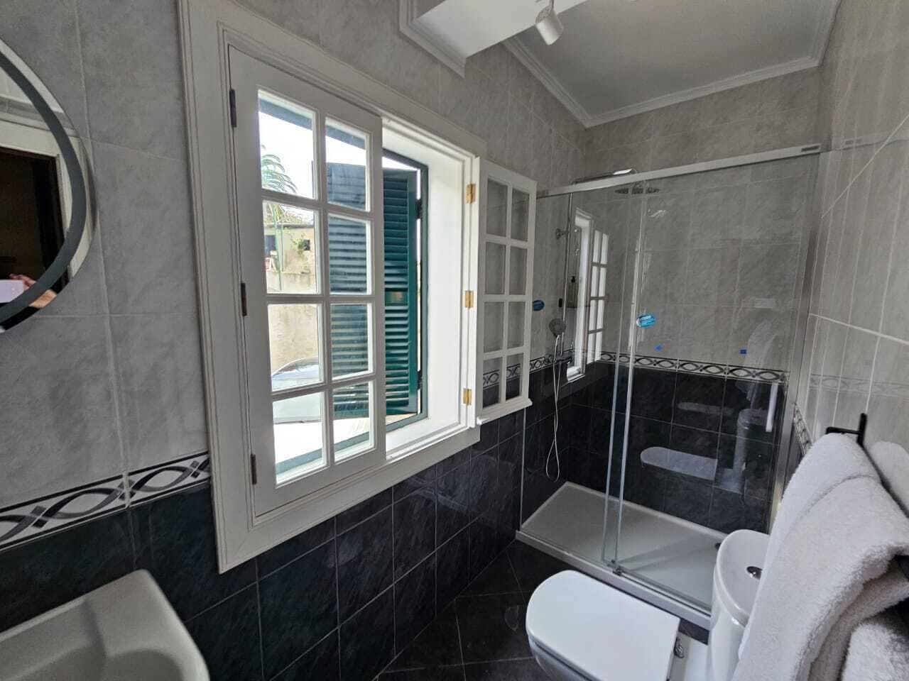 Un baño con ducha, inodoro y ventana con contraventanas verdes.