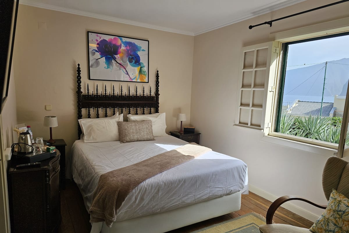 A bedroom with a large bed, a headboard, bedside tables, a chair, a window with a view, and a painting on the wall.