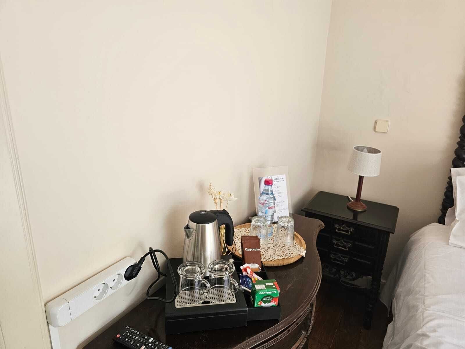 Habitación de hotel con cama, mesita de noche con lámpara, bandeja con botella de agua, cafetera y caja de cappuccino.