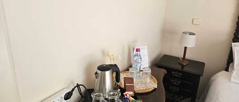 A hotel room with a bed, a bedside table with a lamp, a tray with a water bottle, a coffee maker, and a box of cappuccino.
