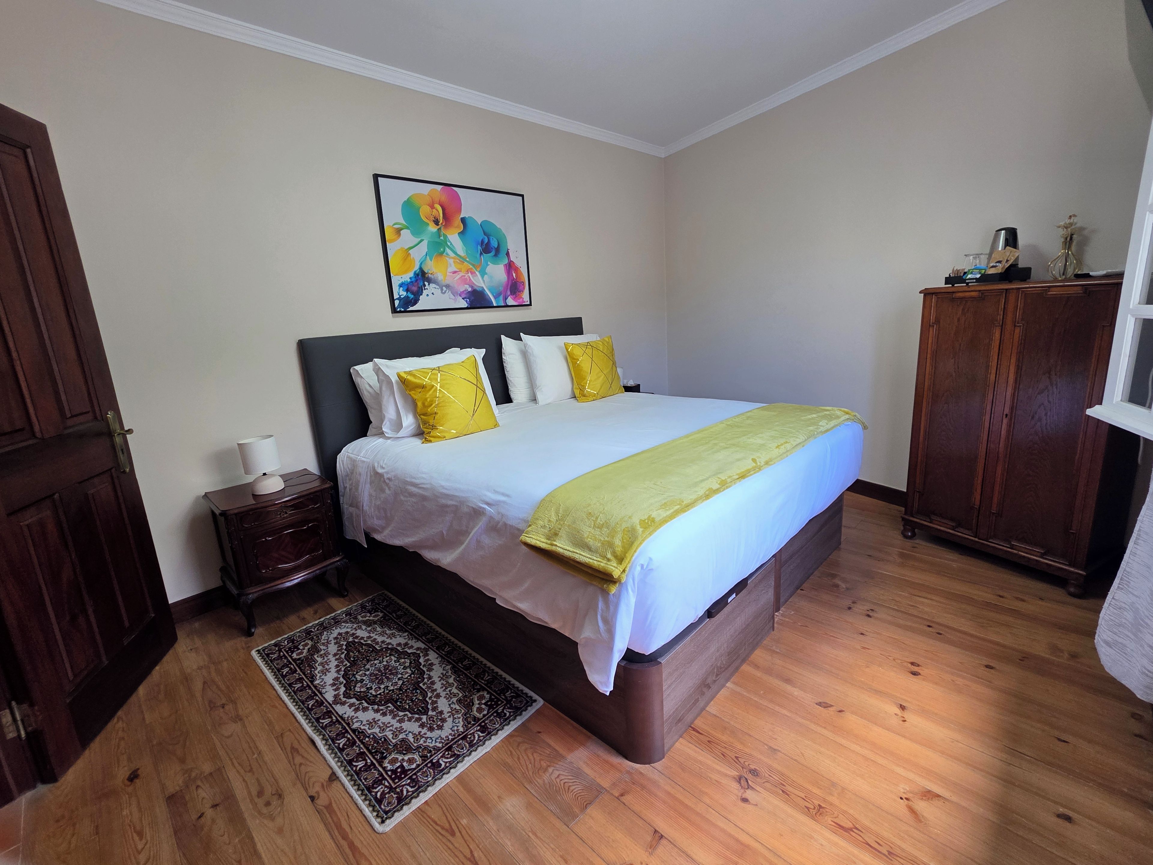 A bedroom with a bed, a nightstand, a wooden wardrobe, a decorative rug, and a painting on the wall.
