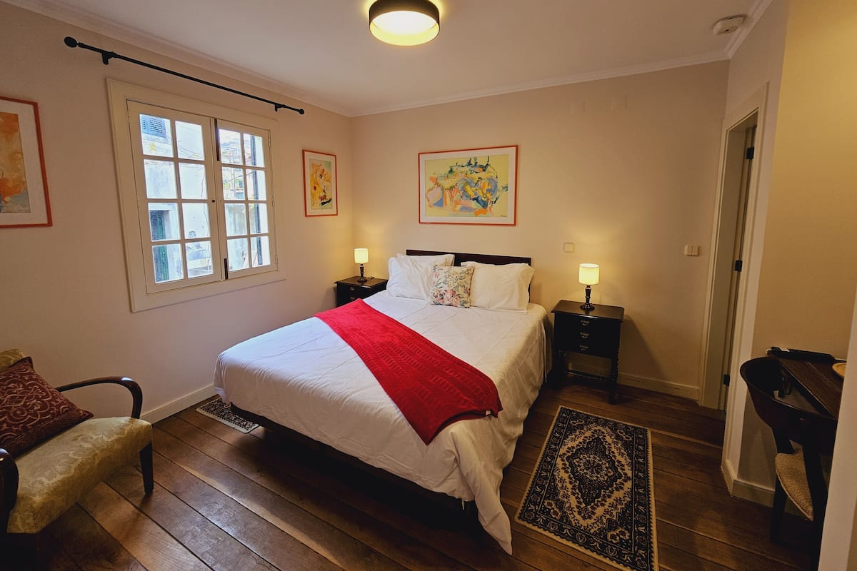 A bedroom with a large bed, two bedside tables, a chair, a window, and artwork on the walls.