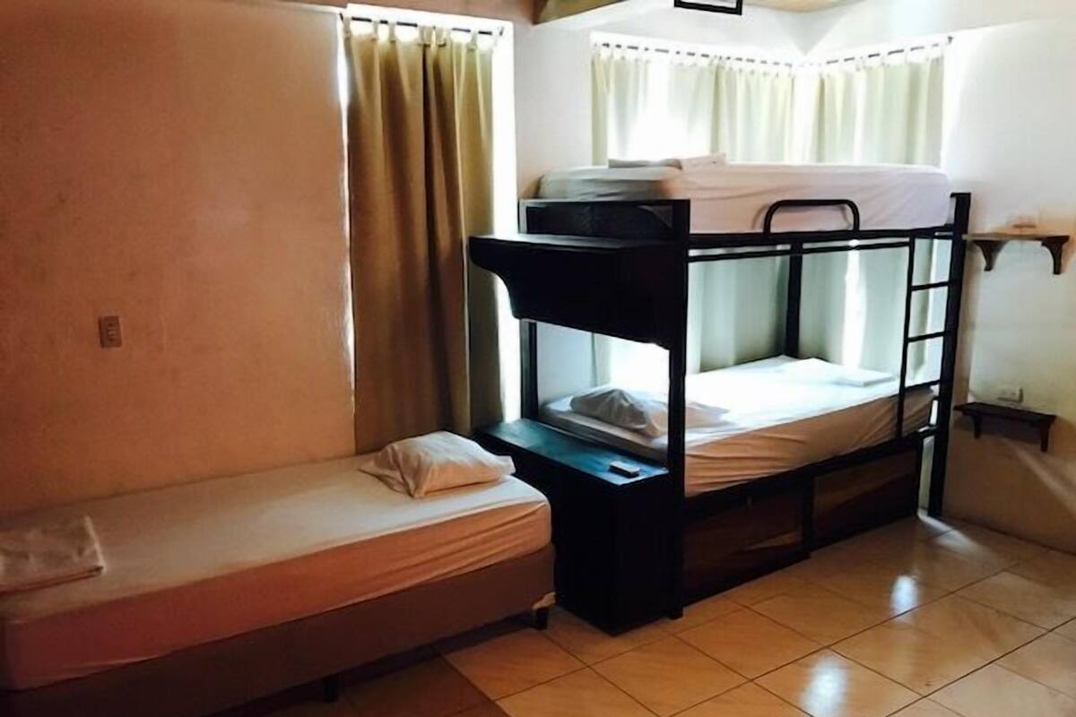 A dormitory room with bunk beds, a small bedside table, and a window with curtains.