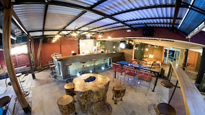 A spacious indoor area with a bar, wooden stools, and a central seating area with a fire pit. - Pachamama - Hotel - Hostel (Managua)
