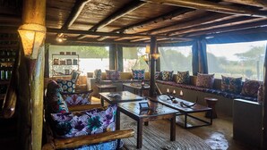 A rustic interior with wooden beams, a sofa with colorful cushions, a coffee table, and a bookshelf. - Ikoma tented Camp (Serengeti)