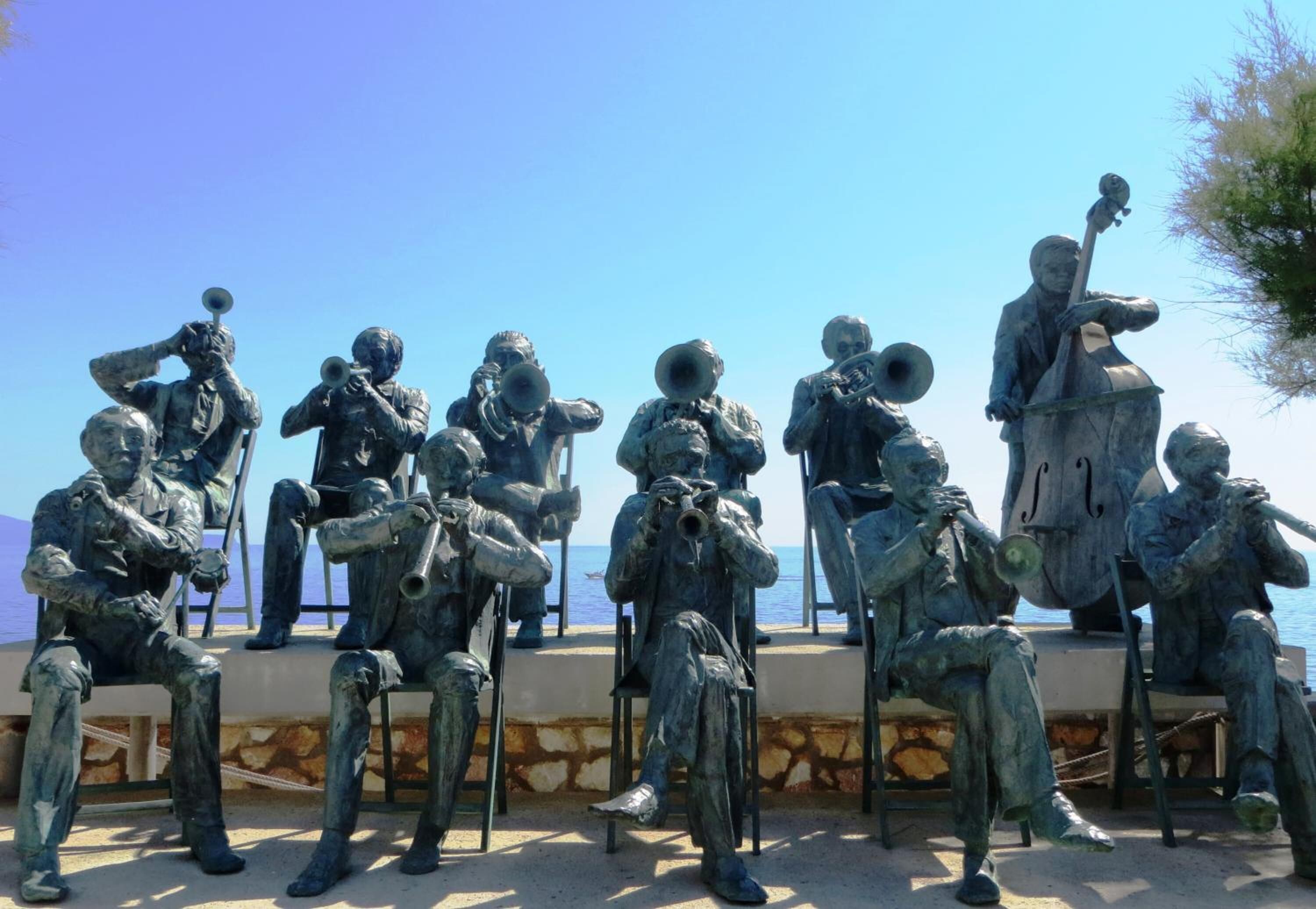 Eine Reihe bronzener Statuen von Musikern, sitzend auf Stühlen, angeordnet auf einer Steinterrasse mit freiem Blick auf das Meer.