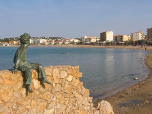 Eine Statue einer sitzenden Figur auf einer Steinmauer am Strand, im Hintergrund sind Gebäude zu sehen.