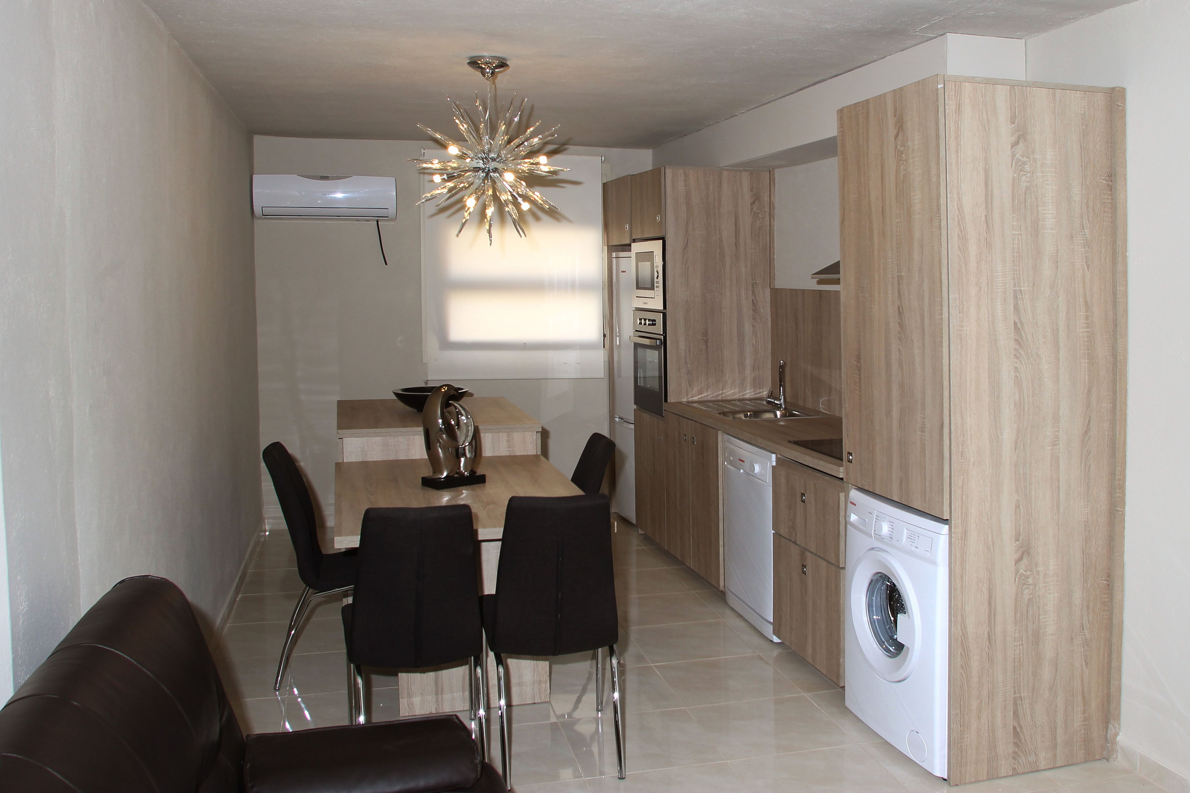 A compact kitchen with a built-in washing machine, a dining area with a table and chairs, and a modern chandelier.